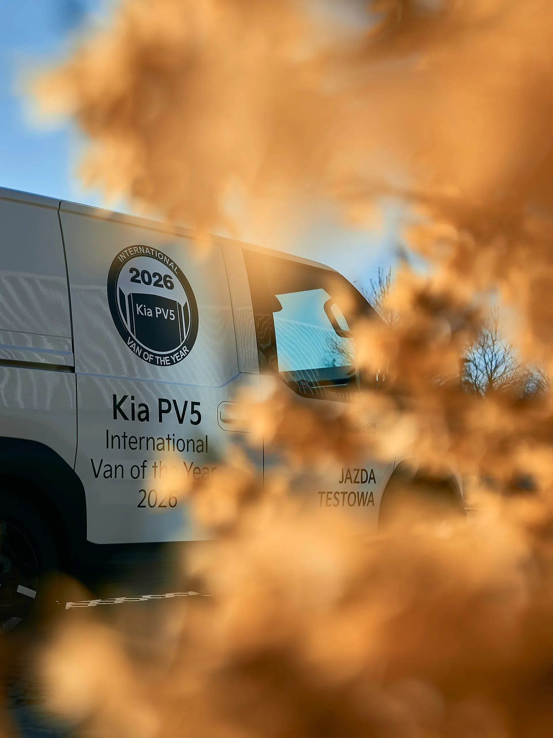 Kia PV5 z naklejką International Van of the Year 2026, sfotografowana przez suche trawy